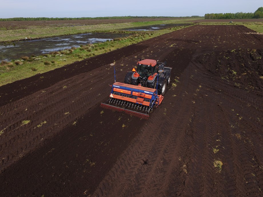 Low angle drone picture, showing a tractor towing machinery across a bare peat field. In the background are two other partially rewetted polders.