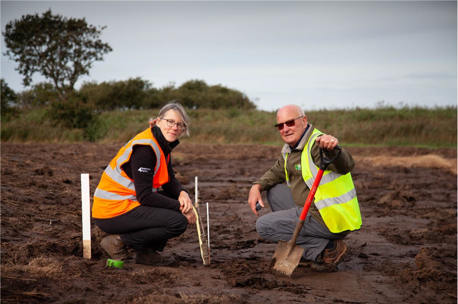 2 people, in Hi Vis vests, planting reeds.