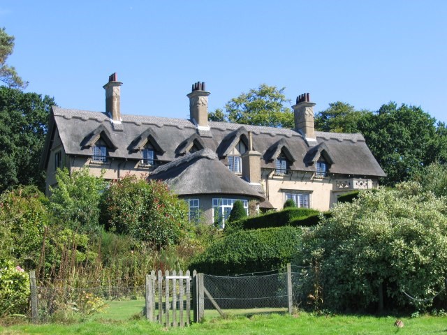 A cream coloured large country house with 3 chimneys and a thatched roof. The roof includes 6 gables, and the house is surrounded by trees.