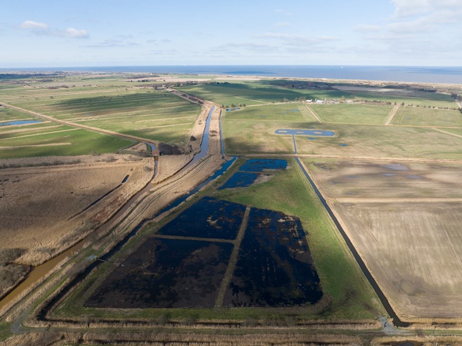 Aerial photograph of the Horsey Wetland Farm. It has five polders filled with water, next to a water channel running through the landscape. The water in the channel is brown. In the background the sea stretches out to the Horizon.